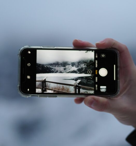 Mão branca segurando o celular e fotogrando uma região da Cracócia, composta por neve, montanha e uma pequena ponte de madeira
