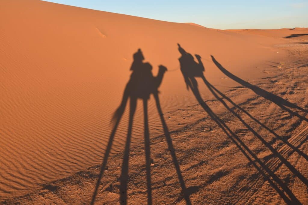 silhuetas de três pessoas no deserto, sendo que duas delas estão montadas em camelos, a areia é bem laranja ao pôr do sol no Deserto do Saara, em Merzouga, que ilustra o post de chip celular Marrocos