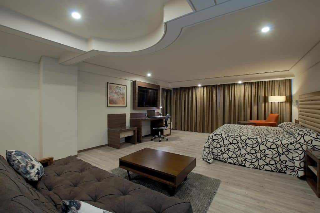 Quarto Deluxe do Econo Express Hotel, de 48 m², com cama de casal, mesa com notebook em cima, cadeira giratória, poltrona laranja e um sofá marrom escuro