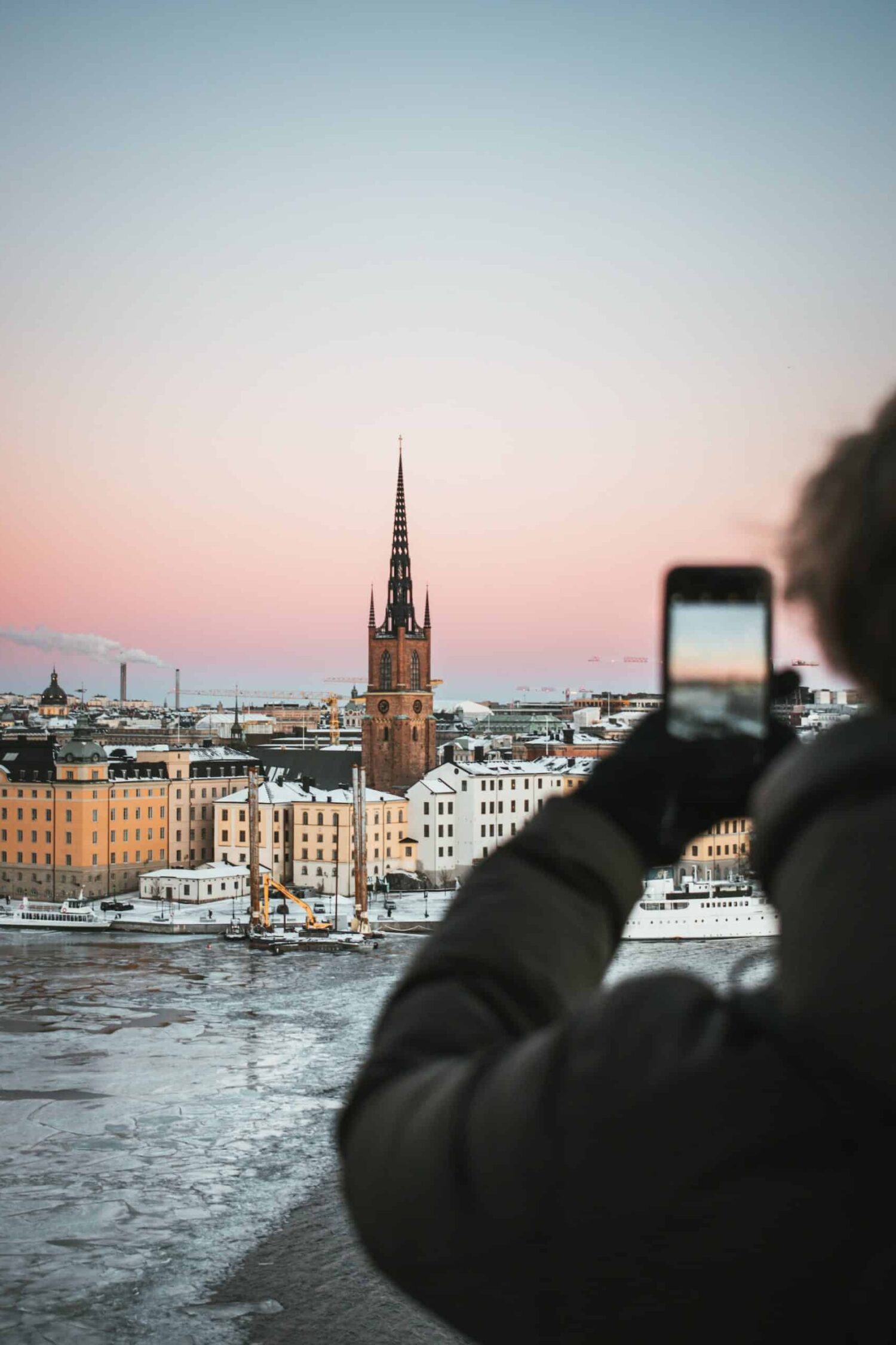Homem usando blusa de frio cinza escuro e luva preta, segurando o celular e fotografando a região de Estocolmo, com prédios históricos, torre e lago congelado