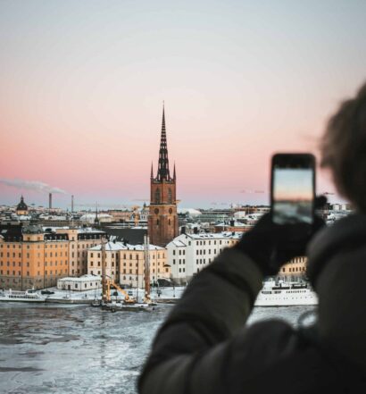 Homem usando blusa de frio cinza escuro e luva preta, segurando o celular e fotografando a região de Estocolmo, com prédios históricos, torre e lago congelado