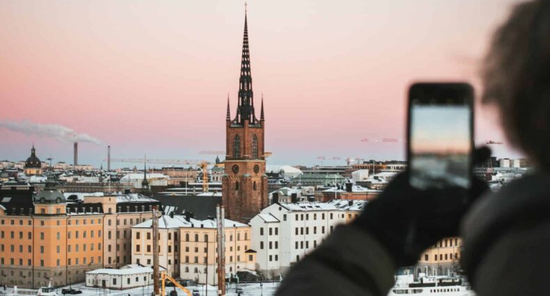 Homem usando blusa de frio cinza escuro e luva preta, segurando o celular e fotografando a região de Estocolmo, com prédios históricos, torre e lago congelado