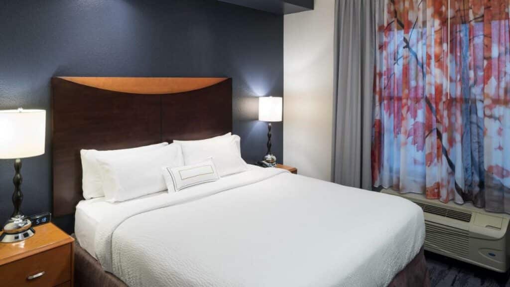 quarto do Fairfield Inn & Suites Orlando at Sea World com cama de casal, com mesinha e luminária de ambos os lados, aquecedor embaixo da janela com cortinas coloridas, sendo um dos hotéis em Orlando