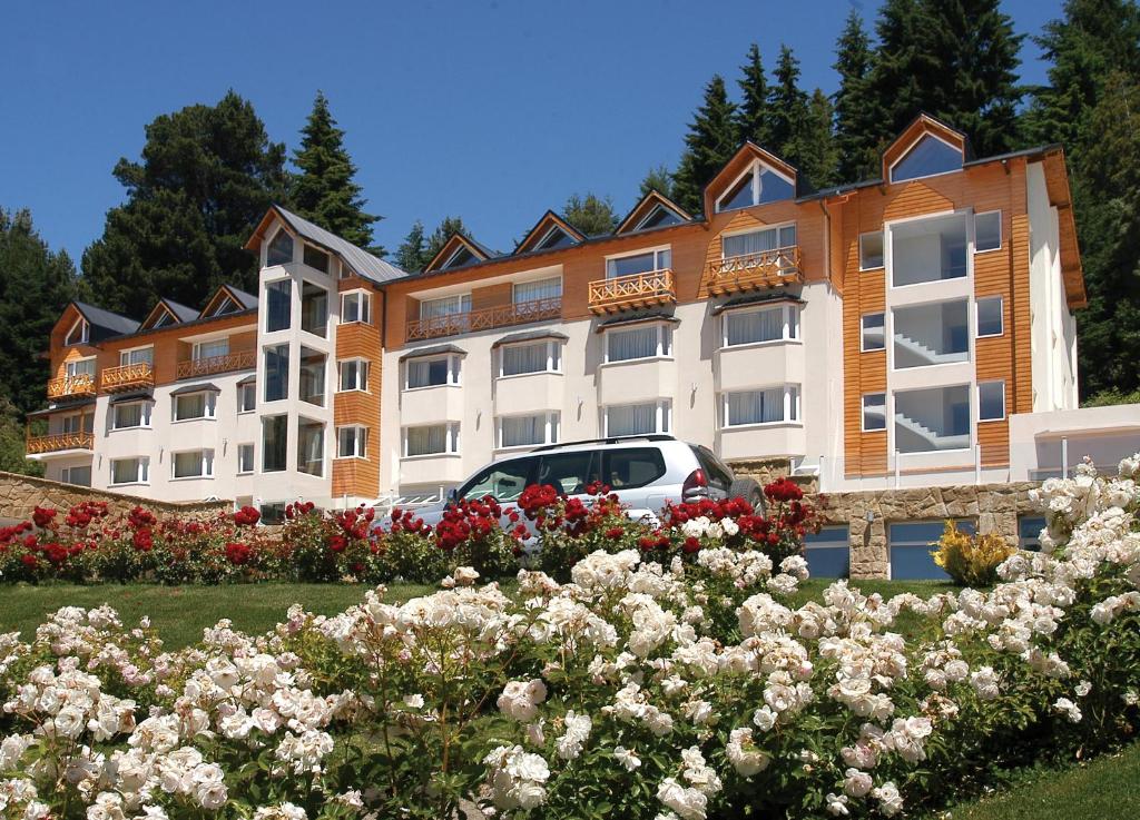 Frente do Huinid Bustillo Hotel & Spa com várias janelas em cores brancas, com um carro cor cinza parada e a frente do hotel jardim com rosas vermelhas e brancas. Representa hotéis em Bariloche.