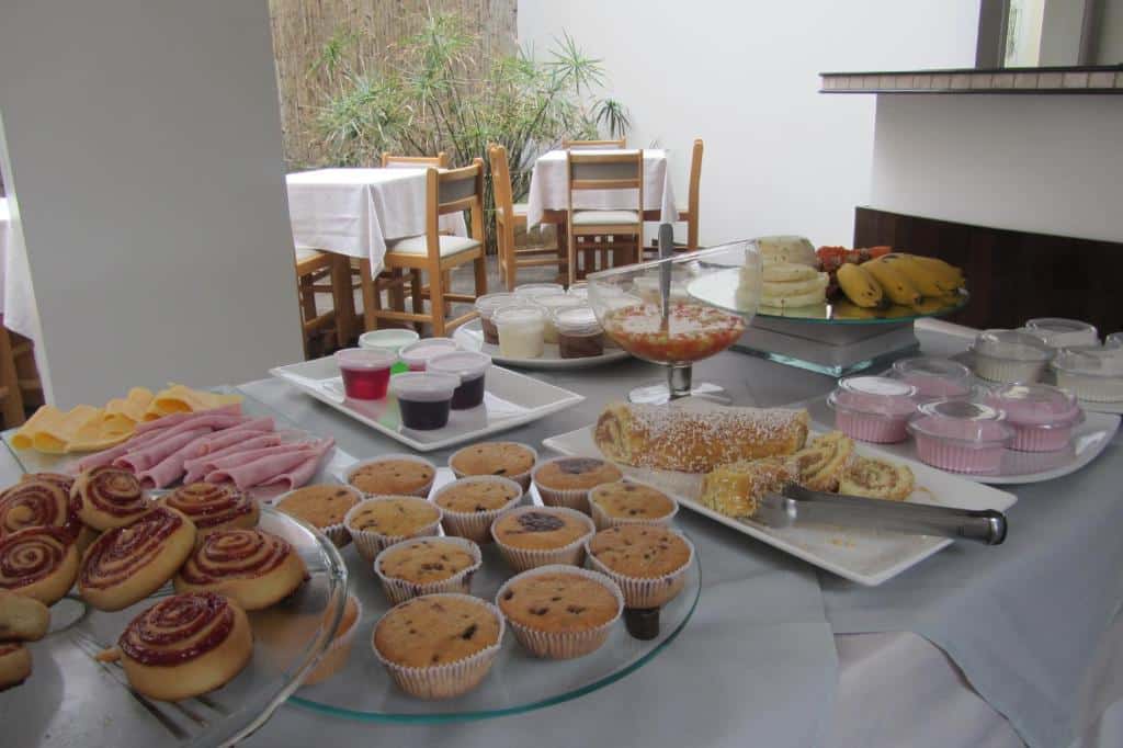 Mesa com opções de café da manhã do Golden Suíte Hotel com pães docas, frios, frutas, e cadeiras e mesas de madeira mais ao fundo
