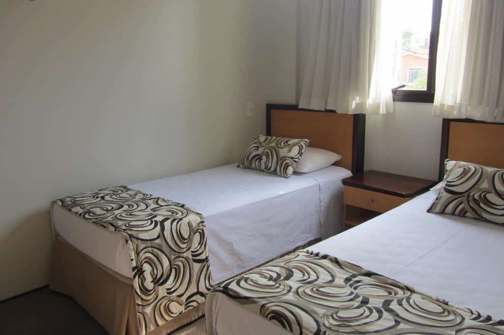 Quarto do Golden Suíte Hotel com duas camas de solteiro, uma janela com cortinas e um pequena mesinha de cabeceira entre as duas camas
