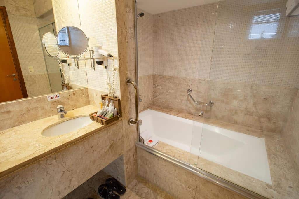 Banheiro do Golden Tower Pinheiros by Fênix Hotéis com uma banheira, uma pia, espelho, secador de cabelo e alguns produtos de higiene sob a pia, para representar hotéis perto do Allianz Parque