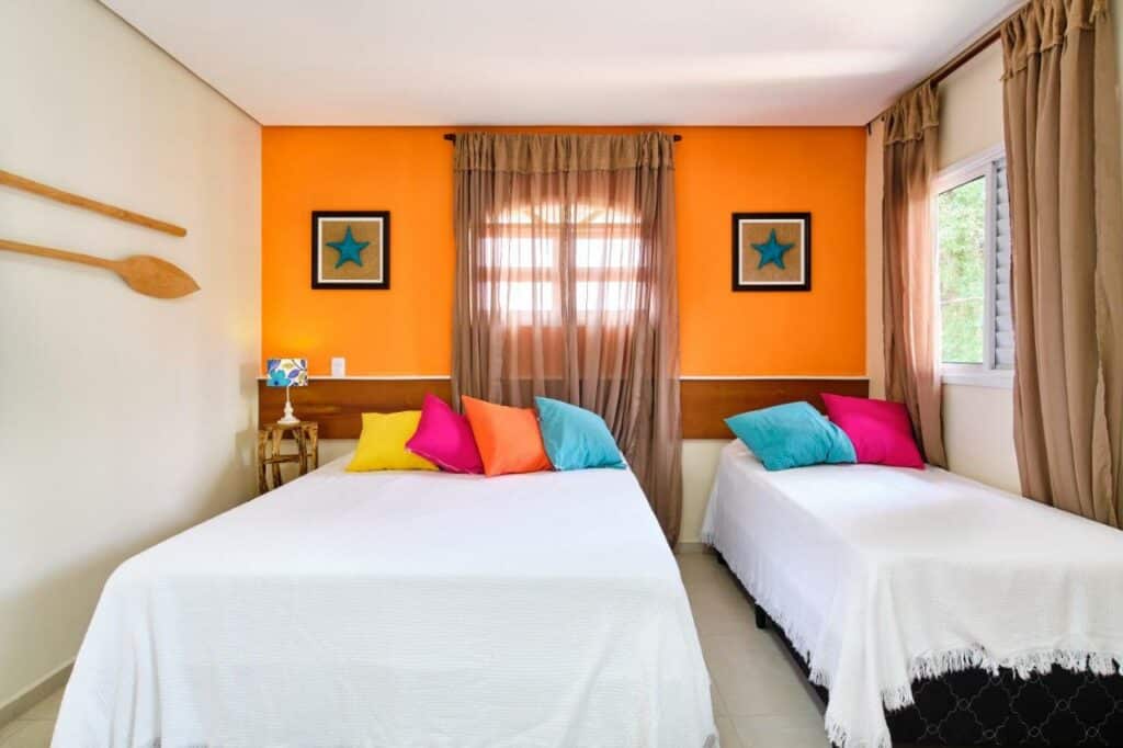 Quarto do Green Haven Residence com uma cama de casal e uma de solteiro, com duas janelas, uma parede laranja com quadros e almofadas coloridas sob as camas