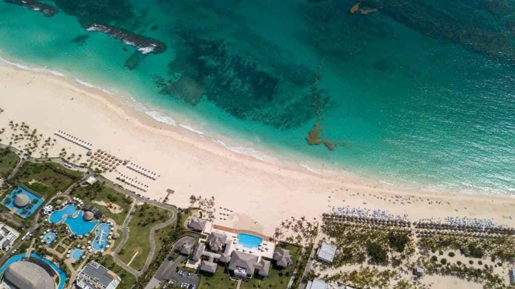 Vista aérea do Hard Rock Punta Cana, sendo metade da imagem mostrando o hotel - com piscinas, teto dos prédios do local e várias espreguiçadeiras e guarda-sóis espalhados na areia branca - e a outra metade da imagem mostrando o mar cristalino azul da ilha