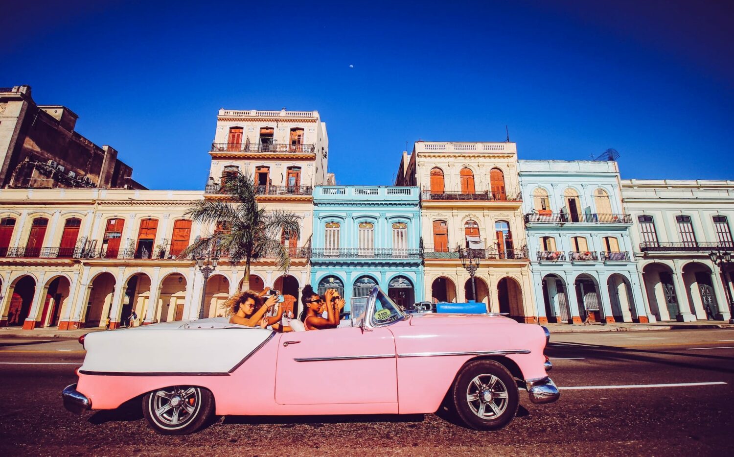 Mulheres andando em um carro antigo conversível rosa, tirando fotos com o celular na mão de uma região de Havana, na Cuba, que tem prédios antigos laranja e azul, com várias janelas, em um dia de céu azul