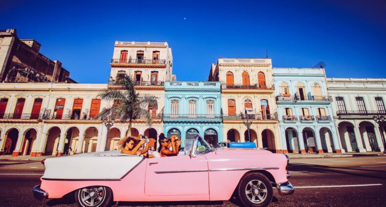 Mulheres andando em um carro antigo conversível rosa, tirando fotos com o celular na mão de uma região de Havana, na Cuba, que tem prédios antigos laranja e azul, com várias janelas, em um dia de céu azul