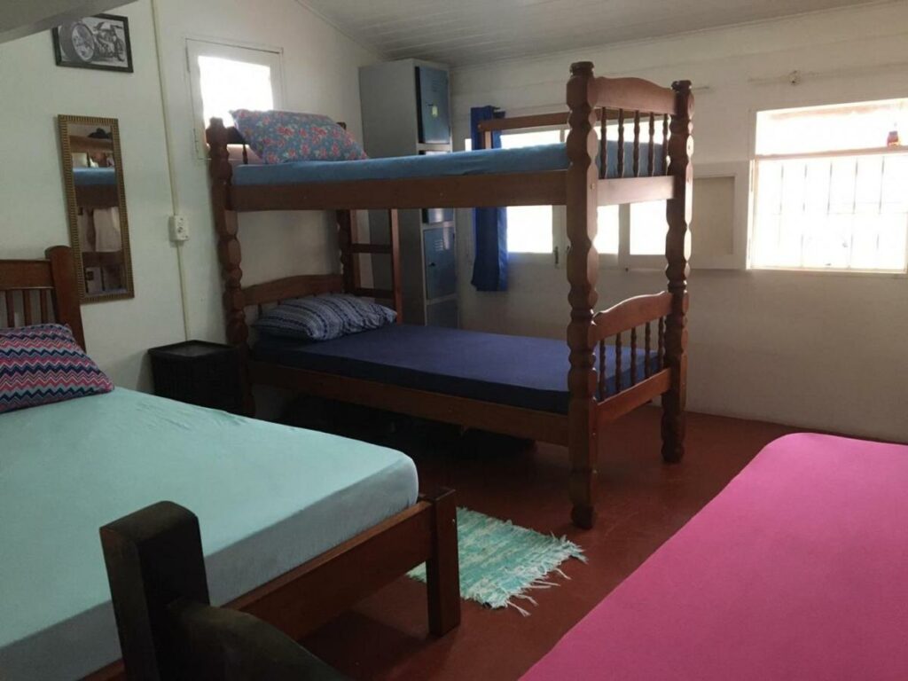 Uma quarto compartilhado no Hostel 7 praias com uma beliche e duas camas de casal, além de alguns armários com chave e duas janelas com cortinas, para representar hostels em Ubatuba
