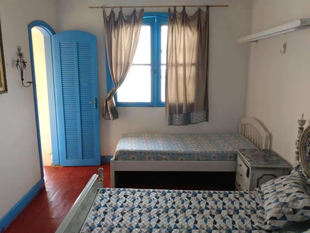 Quarto no Hostel Mama House com duas camas de solteiro, uma janela com cortinas, e uma pequena mesinha de cabeceira entre as camas