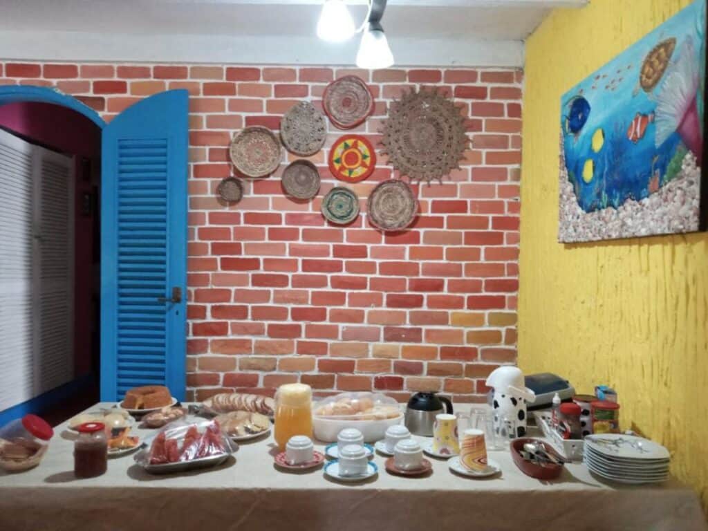 Mesa para refeições no Hostel Mama House com xícaras, pães, frutas e pratos a disposição dos hóspedes