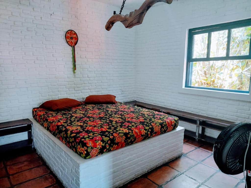 Um quarto no Hostel Terra das Tribos com uma cama de casal, uma janela do lado direito e um ventilador de chão