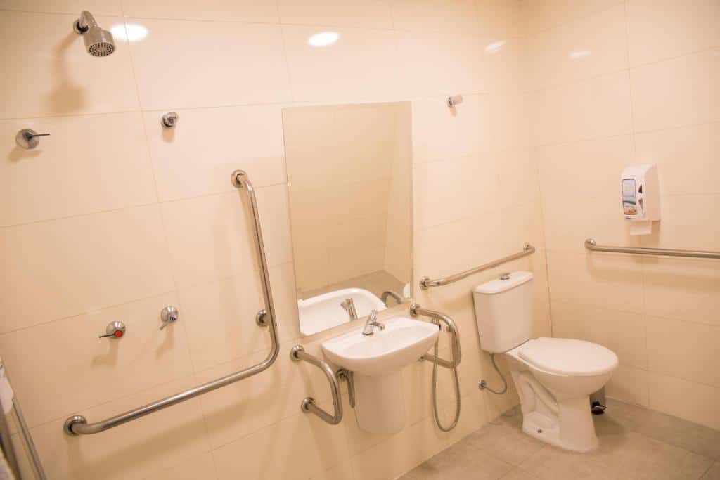 Banheiro adaptado com barras de apoio, uma pia, um vaso sanitário e um espelho, além do chuveiro, no Hotel Aero Sleep Campinas