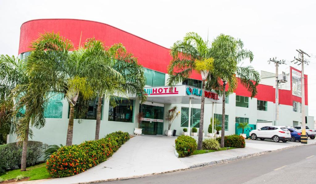 Fachada do Hotel Alji, um prédio comercial em tons de vermelho e branco, com coqueiros e alguns arbustos na frente, além de vagas abertas para carros