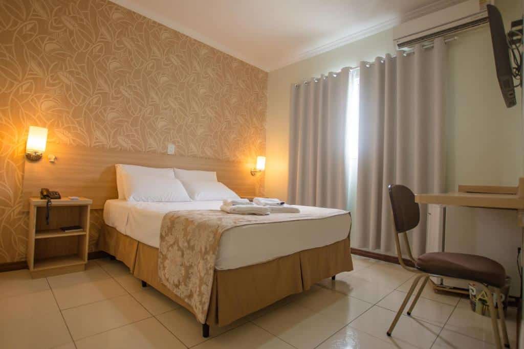 Quarto no Hotel Alji com uma cama de casal, duas luminárias presas na cabeceira, uma televisão, uma bancada com cadeira e um frigobar, para representar hotéis perto do Aeroporto de Viracopos em Campinas