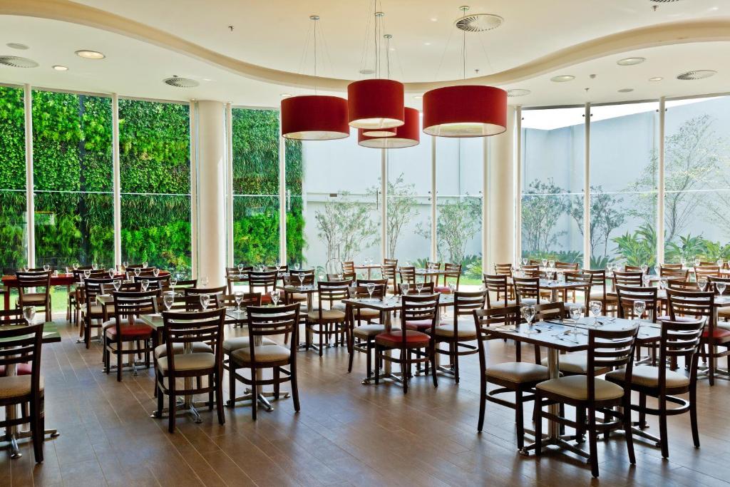 Área de refeições do Hotel Panamby São Paulo com paredes de vidro que dão vista para o jardim, do lado de dentro há mesas e cadeiras de madeira