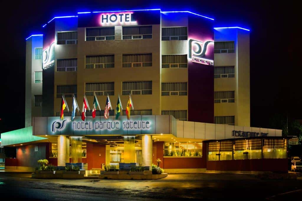 Frente iluminada do Hotel Parque Satelite, durante uma noite, com bandeira de países na entrada e vários letreiros iluminados espalhados pela fachada escrito "hotel parque satelite", "restaurante" e "hotel"