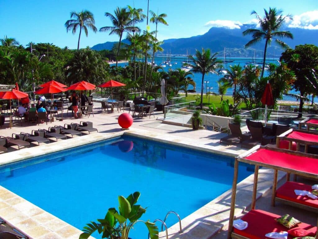 Piscina do Hotel Itapemar - Ilhabela com vista para a praia e, ao redor, da piscina há um deck com espreguiçadeiras vermelhas e toalhas
