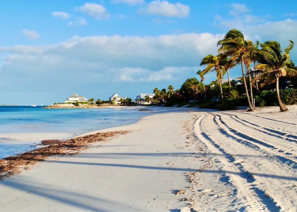 vista da praia de areias clarinhas das Ilhas Berry, em Bahamas, com mar azul claro, quase cor do céu com nuvens e, mais ao fundo, algumas construções e palmeiras