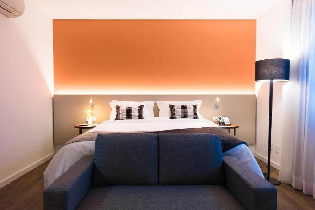 Quarto do Intercity São Paulo Anhembi com uma cama de casal e um sofá de dois lugares na frente da cama, o chão é de madeira, a parede atrás da cama tem iluminação indireta, do lado direito há um abajur de chão e do lado esquerdo um ar-condicionado no teto