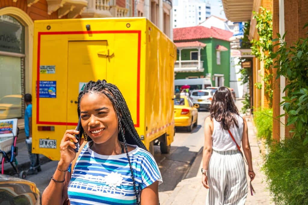 Uma moça negra com tranças no cabelo segurando um celular no ouvido enquanto anda em uma rua cheia de carros em Cartagena