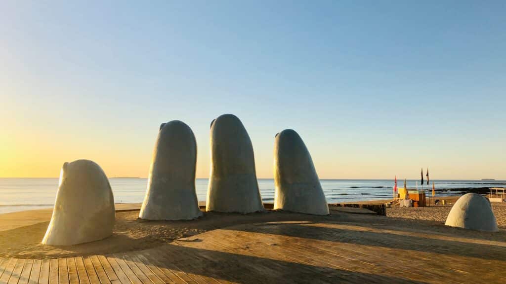 Vista do monumento La Mano, em Punta del Este, com pontas dos dedos saindo do chão com vista para o mar no pôr-do-sol. Essa foto ilustra o post sobre seguro viagem Uruguai.