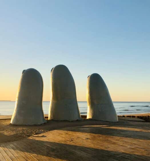Vista do monumento La Mano, Punta del Este, com pontas dos dedos saindo do chão com vista para o mar no pôr-do-sol.