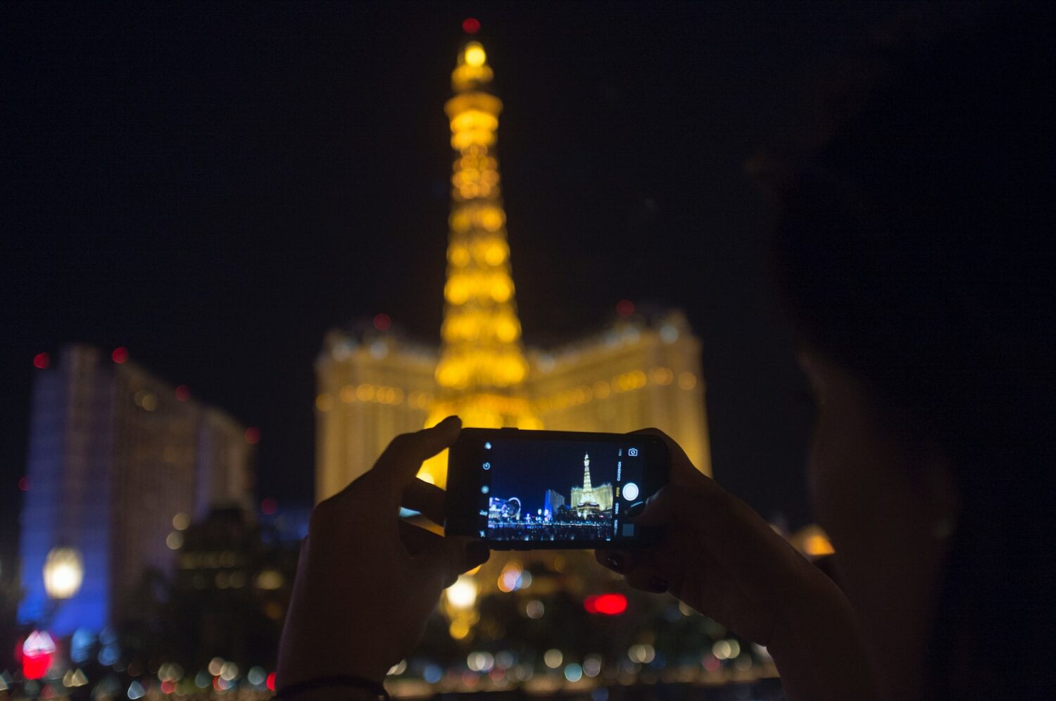 Foto de silhueta de pessoa tirando uma foto com o celular da Torre Eiffel de Las Vegas, iluminada ao fundo na parte da noite.