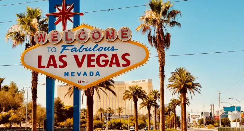 A placa "Bem-vindo à fabulosa Las Vegas" fica cercada por palmeiras em um dia ensolarado, com a paisagem urbana ao fundo.