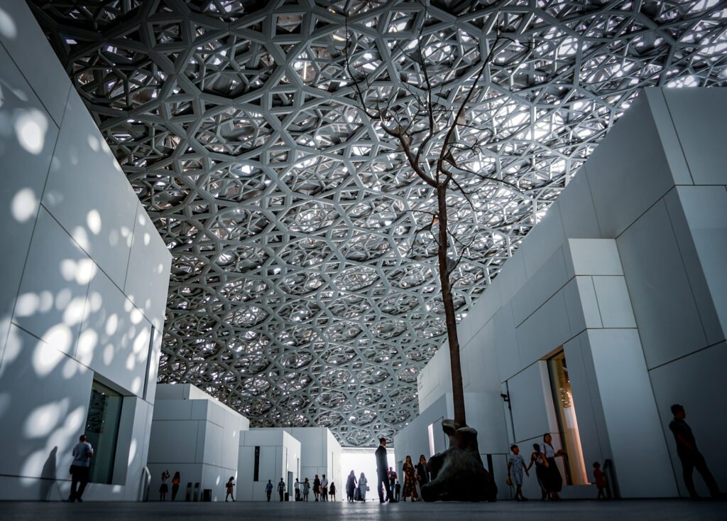 Pessoas caminhando entre as paredes brancas do museu para ilustrar o post sobre chip de celular para Abu Dhabi. O teto é coberto por uma estrutura de ferro abrangente. - Foto: Agnieszka Kowalczyk via Unsplash