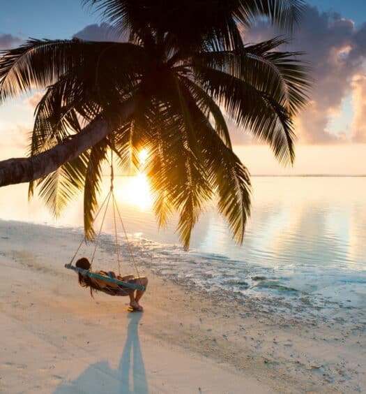 pessoa relaxando em um balanço pendurado em um coqueiro que se projeta para frente, em direção da praia de águas azuis cristalinas e areia branca nas Maldivas