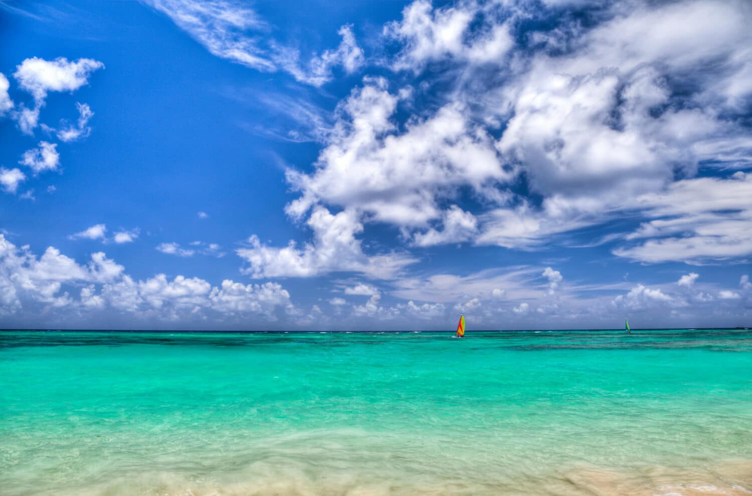 Mar cristalino azul, com um barco de vela nas cores verde florescente, vermelho e laranja, e um céu azulado com nuvens brancas