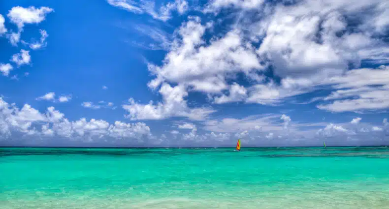 Mar cristalino azul, com um barco de vela nas cores verde florescente, vermelho e laranja, e um céu azulado com nuvens brancas