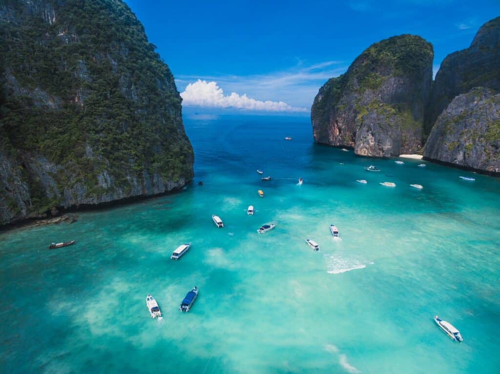 Barcos e lanchas em água cristalina em alto mar emoldurado por duas rochas com vegetação, céu azul com algumas nuvens brancas para representar o seguro viagem Phuket.