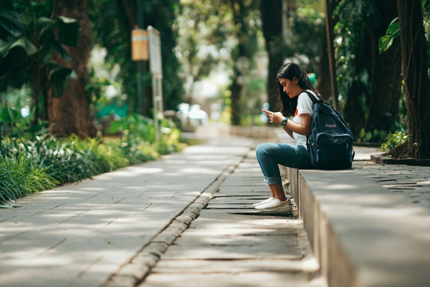 Menina usando calça jeans e blusa branca, com mochila nas costas, sentada e olhando no celular. O lugar é rodeado de vegetação e a imagem é para ilustrar o post de chip celular Indonésia