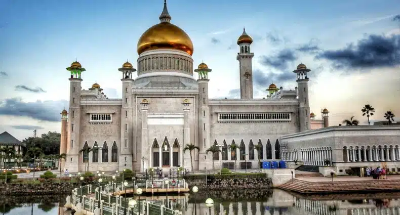 Vista da Grande Mesquita de Omar Ali Saifuddin, Brunei de cor branca, teto dourado, com várias janelas em frente ao lago durante o dia. Representa chip celular Brunei
