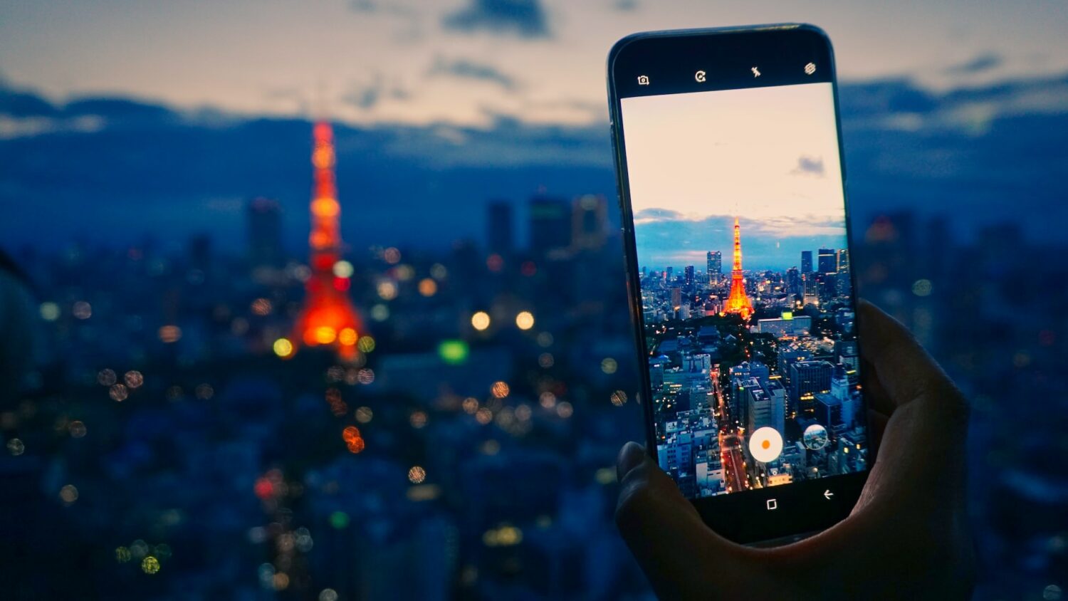 Um celular sendo usado para fotografar a cidade de Tóqui com foco na Torre de Tóquio, que se parece um pouco com a Torre Eiffel