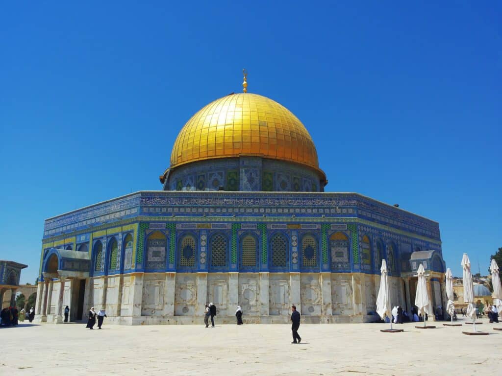 Construção bege com detalhes azuis e verdes e um domo amarelo no topo para ilustrar o seguro viagem Palestina. O céu está azul durante o dia e há pessoas caminhando em frente à mesquita. - Our Tayeh via Unsplash