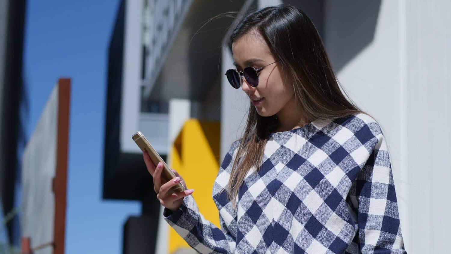 Uma mulher branca de cabelos lisos e castanhos usando um óculso de sol e uma camiseta xadrez segura e olha para um celular em sua mão