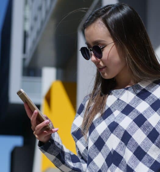 Uma mulher branca de cabelos lisos e castanhos usando um óculso de sol e uma camiseta xadrez segura e olha para um celular em sua mão