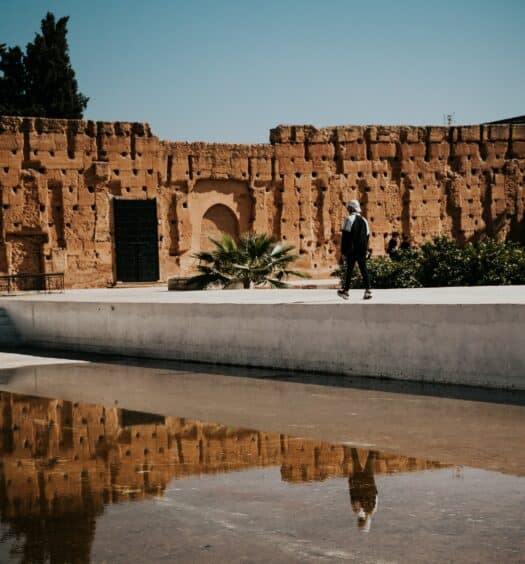 homem andando em uma passarela que leva até as ruínas de pedra em tons de terra e areia do Palácio El Badi, em Marrakesh, para ilustrar o post de chip celular Marrocos