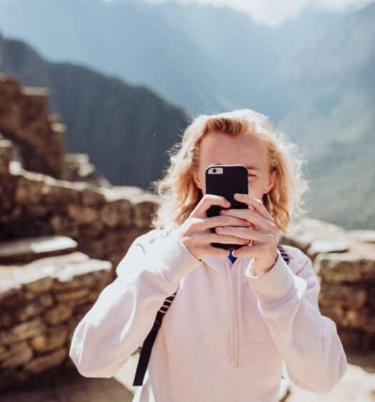 Um jovem branco de cabelo loiro segurando um celular com as ruínas de Machu Picchu ao fundo