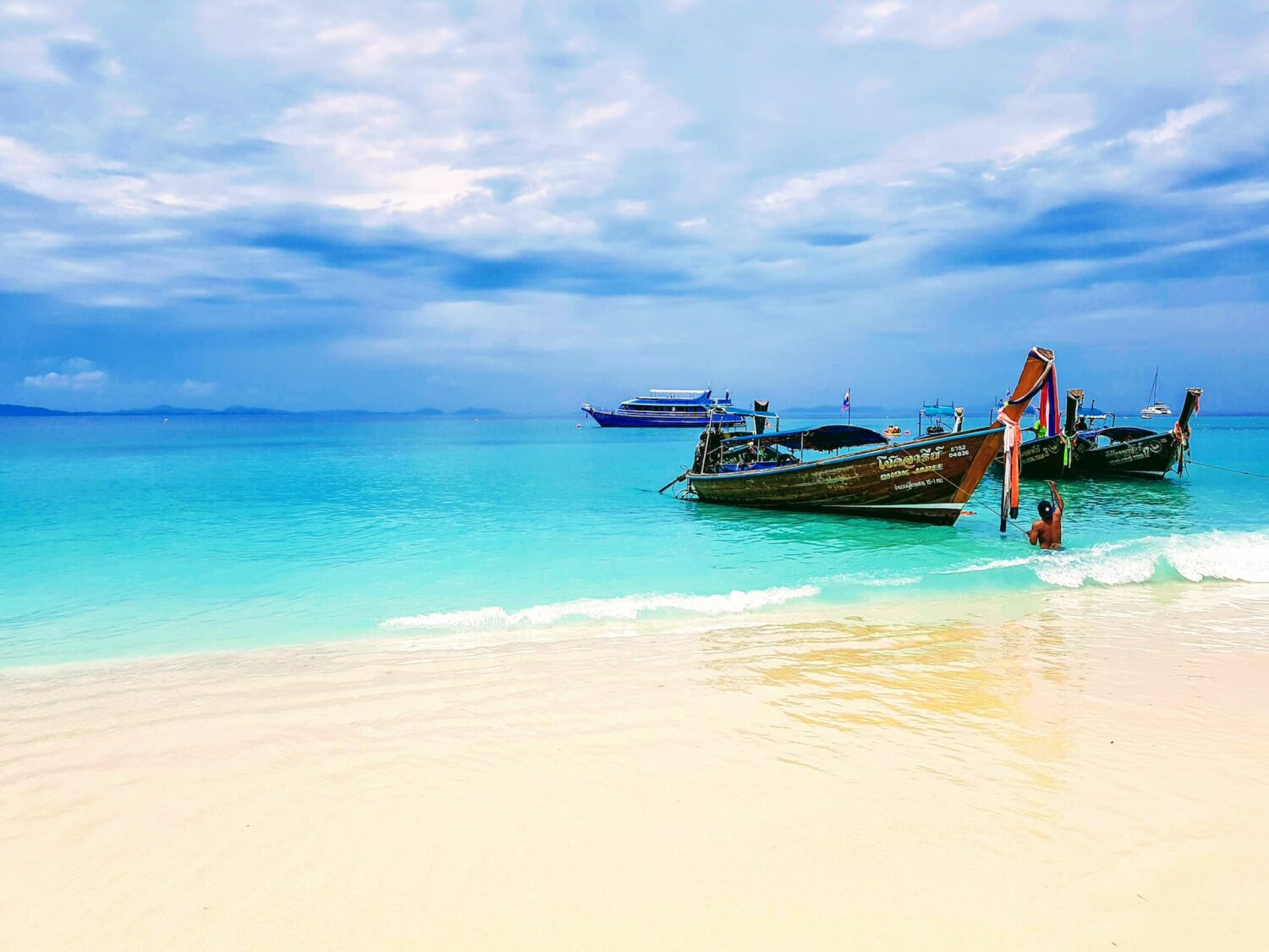 Praia com águas cristalinas em Phuket, com uma faixa de areia branca, ao lado no mar uma canoa, céu azul com algumas nuvens.