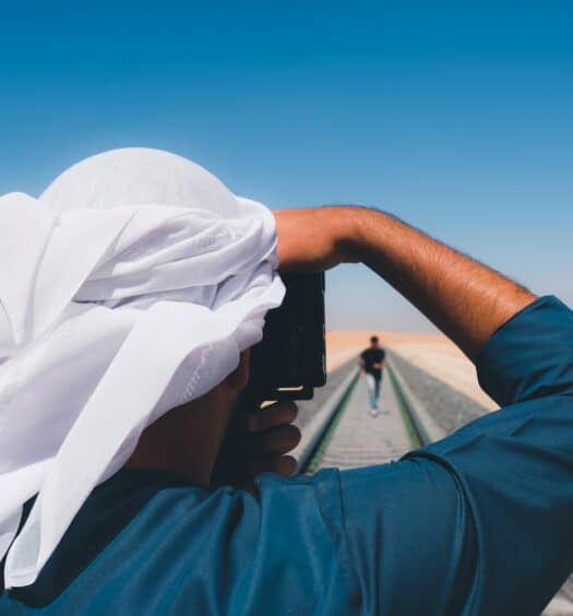 Pessoa de blusa azul de mangas compridas e hijab branco tirando foto de alguém no meio do deserto. - Foto: David Suaza via Unsplash