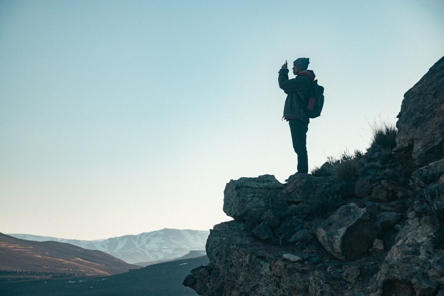 Homem de roupas de inverno em cima de uma rocha e tirando foto com o celular. Atrás dele, o céu é azul e há montanhas na base da imagem. - Foto: Gustavo Zambelli via Unsplash