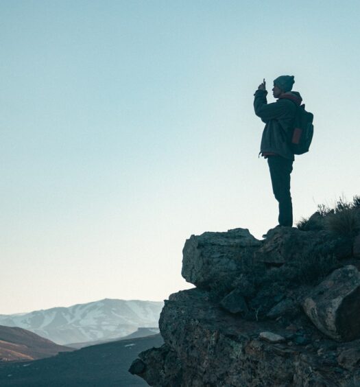 Homem de roupas de inverno em cima de uma rocha e tirando foto com o celular. Atrás dele, o céu é azul e há montanhas na base da imagem. - Foto: Gustavo Zambelli via Unsplash