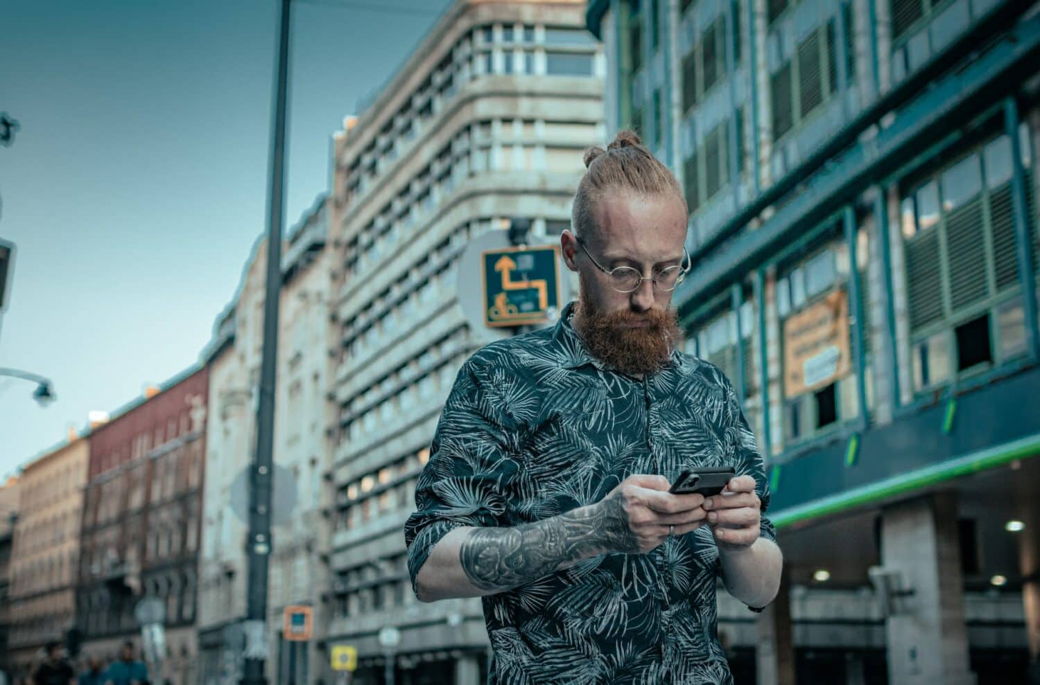 Homem de barba ruiva e óculos mexendo no celular com prédios ao fundo. Ele veste uma camisa de mangas curtas com estampa de folhas verdes e tem uma tatuagem no braço. - Foto: Kat Sazonova via Unsplash